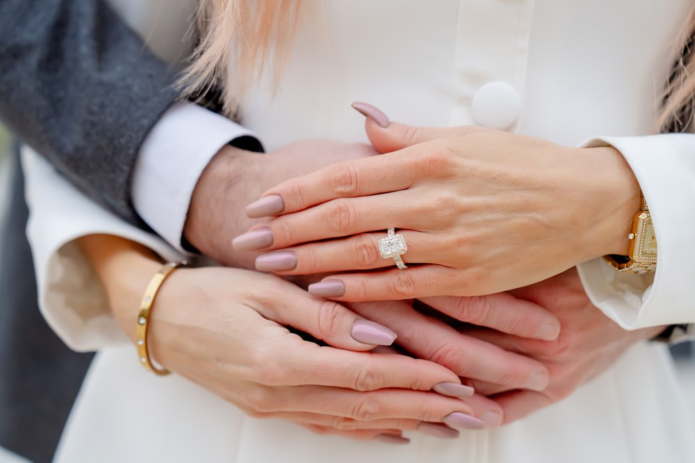 Close-up of a couple's hands, his arms around her white coat and her engagement ring in focus, engagement photo by Alex in Paris