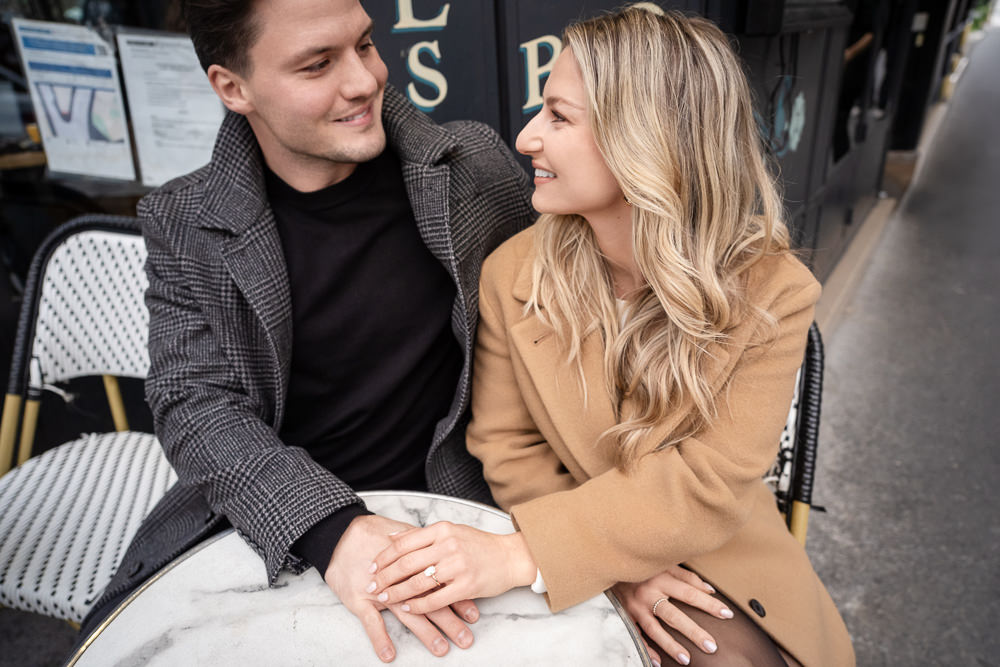 Newly engaged couple holding hands on a marble café table in Paris, her engagement ring in view, photographed by Olga