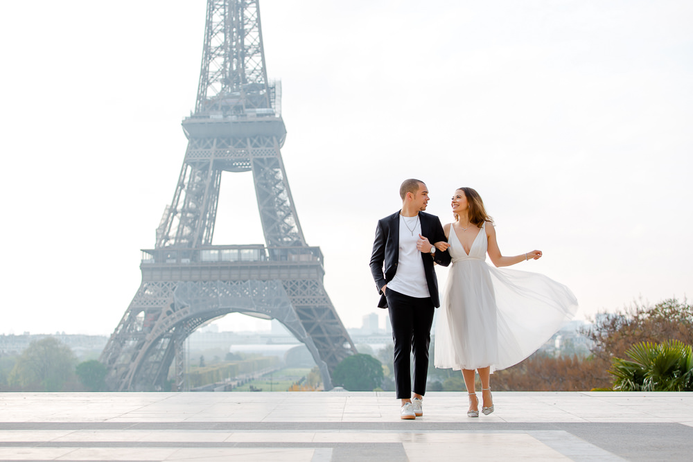 Couple walk arm in arm across the Trocadéro terrace, her white dress in motion, Eiffel Tower behind them, photographed by Alex in Paris