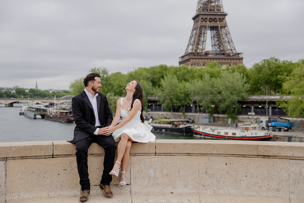 Couple laughing on a stone wall above the Seine with the Eiffel Tower behind, a candid moment by Lena, couples photographer in Paris
