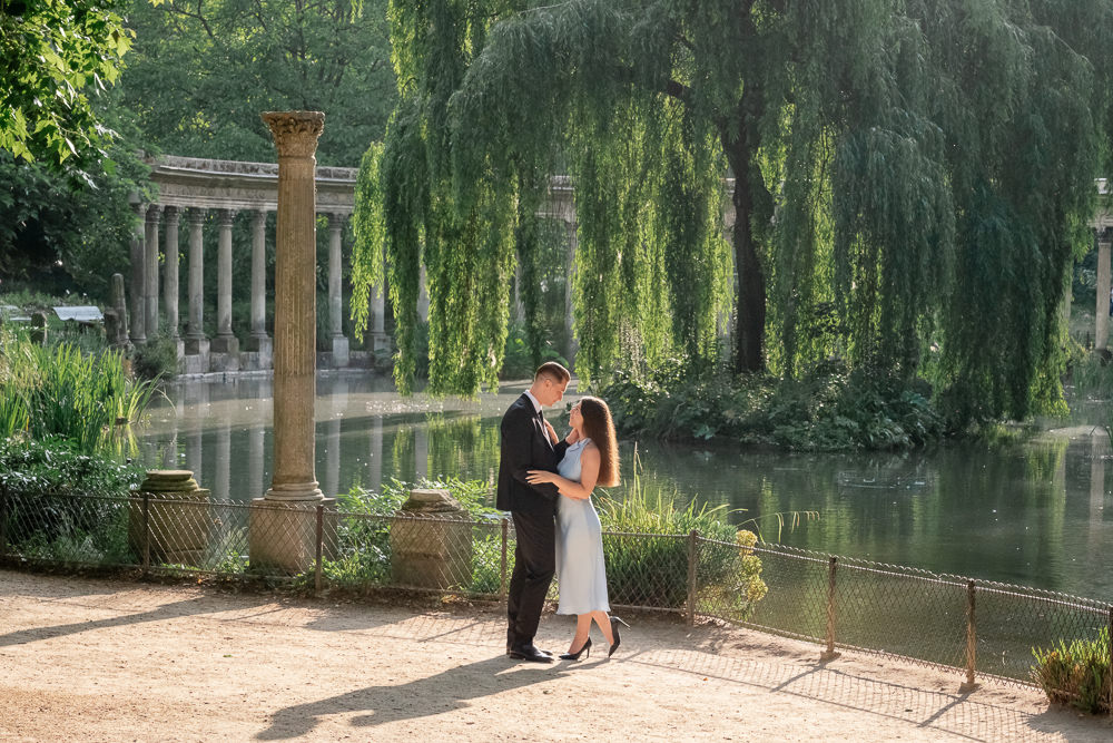 Engaged couple embracing beside the colonnade pond in Parc Monceau at golden hour, photographed by Olga