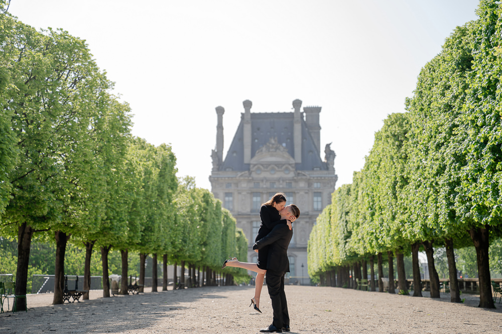 Man lifting his partner for a kiss in a tree-lined alley of the Tuileries Garden, with the Louvre behind them