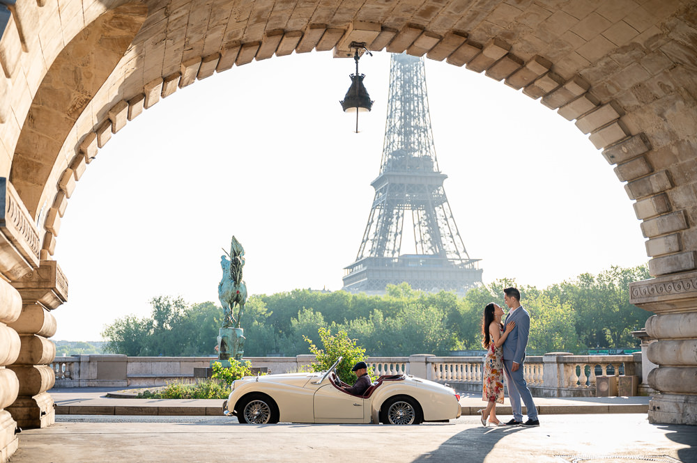 Couple embracing under the stone arch of Bir-Hakeim bridge beside a cream vintage convertible, Eiffel Tower framed behind them